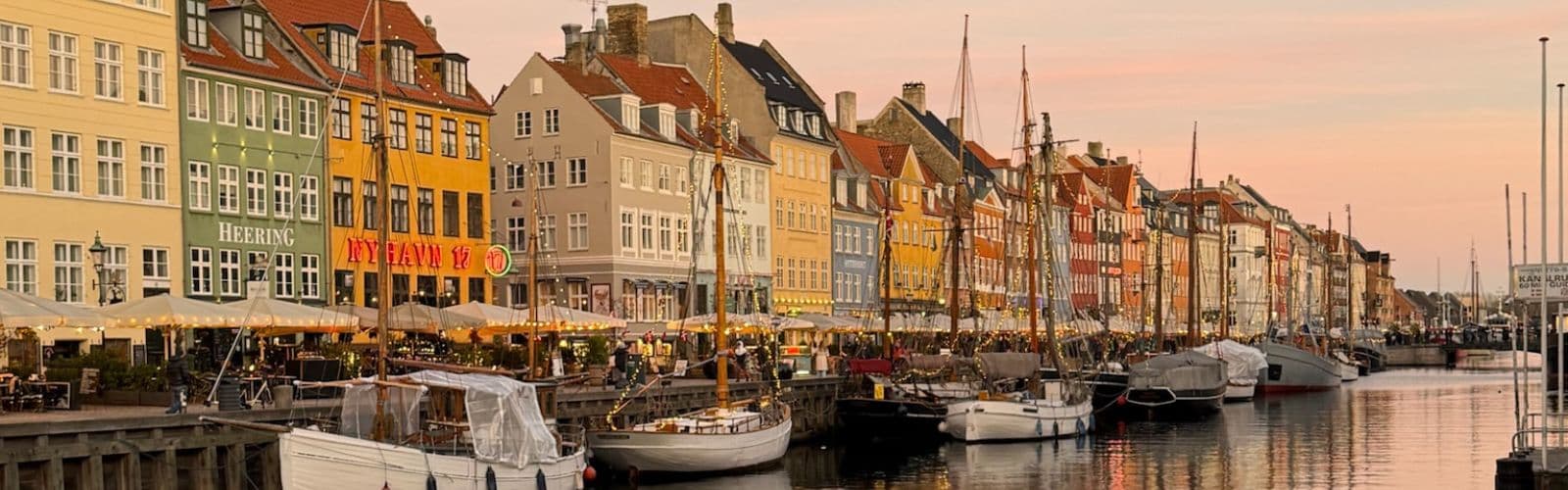 Colorful, narrow houses sit against a harbor filled with boats as the sun sets in the background.